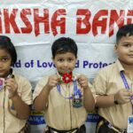 Three smiling schoolchildren in beige uniforms display handmade flower pins on lanyards in front of a banner